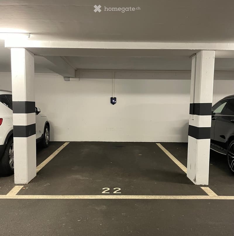 Underground parking space with two cars parked, marked with number 22, white pillars, and a security camera.