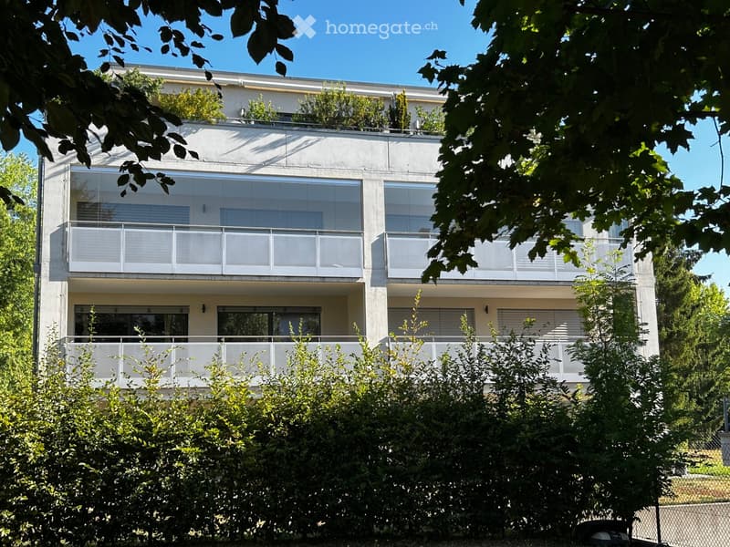 Modern two-story building with large windows and balconies, surrounded by greenery.
