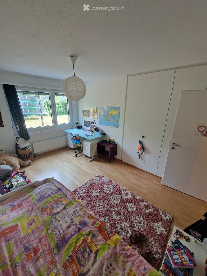 A well-lit bedroom with a colorful bedspread, a wooden floor, a desk with a chair, a wardrobe, a hanging lamp, and various decorations including a world map and picture frames.