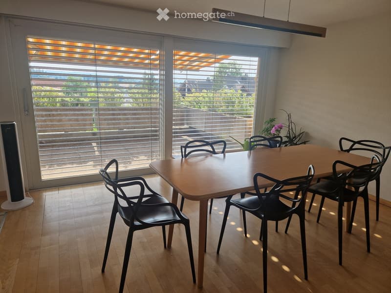 Bright dining area with wooden table, black chairs, and a fan near the window with blinds.