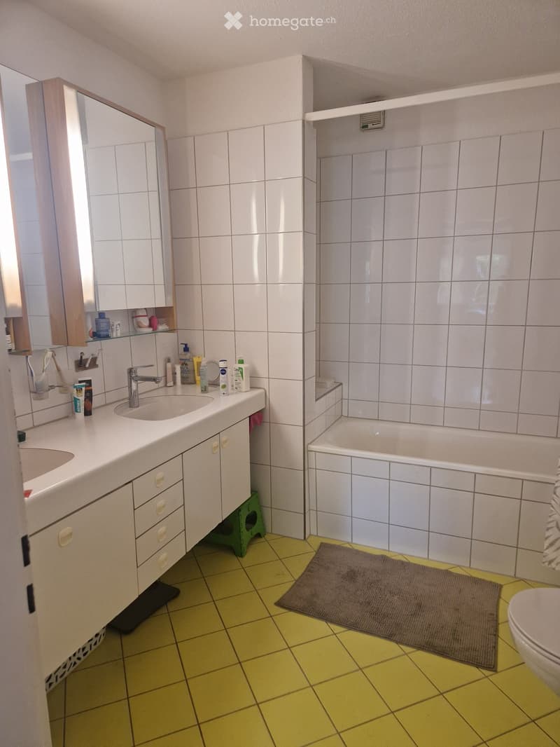 Bright bathroom with yellow tiled floor, white tiled walls, a double sink vanity with cabinets, a bathtub, a mirror, and a toilet. The space includes a towel on the floor and various toiletries on the sink counter.