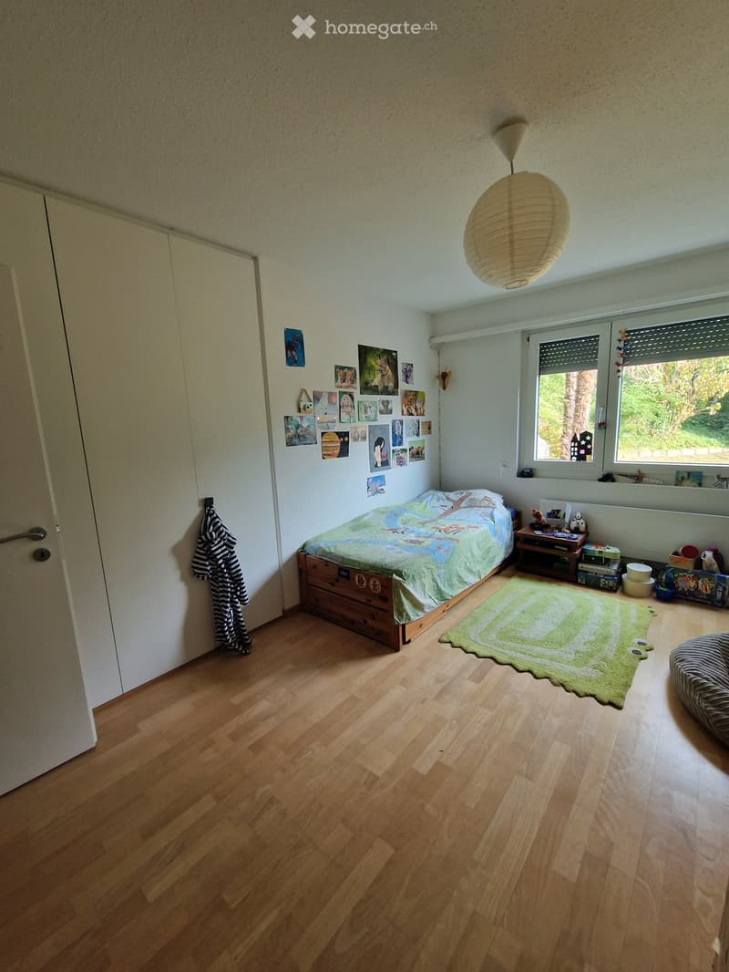 Cozy bedroom with wooden flooring, a single bed with a colorful duvet, a nightstand with a lamp, a green rug, and several picture frames on the wall.