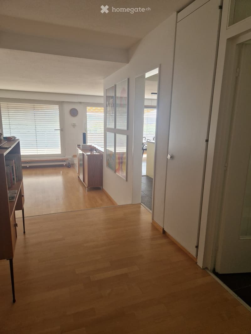 Bright hallway with wooden flooring, large windows, and a view of adjacent rooms. Features a clock on the wall and a small cabinet with books and decorative items.