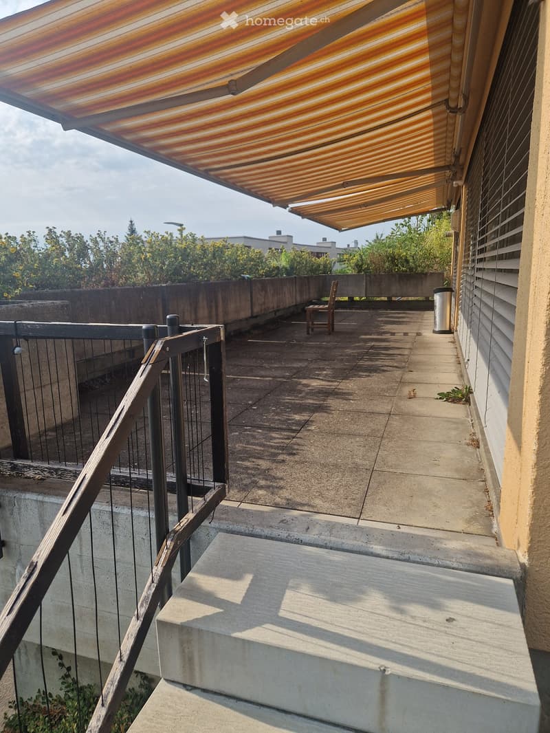 Balcony with a striped awning, concrete floor, wooden railing, and a single wooden chair.