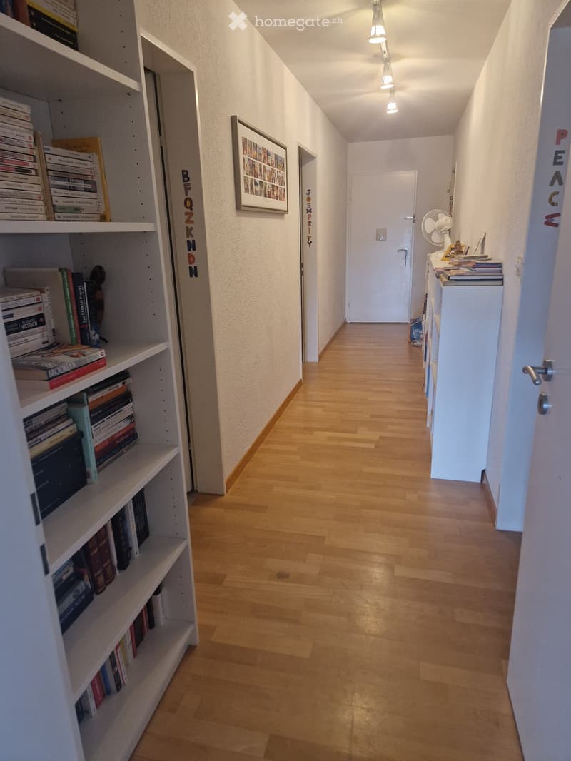 A narrow hallway with wooden flooring, white walls, and a bookshelf filled with books on the left side. A framed picture hangs on the wall, and a small table with a fan and books is visible on the right.