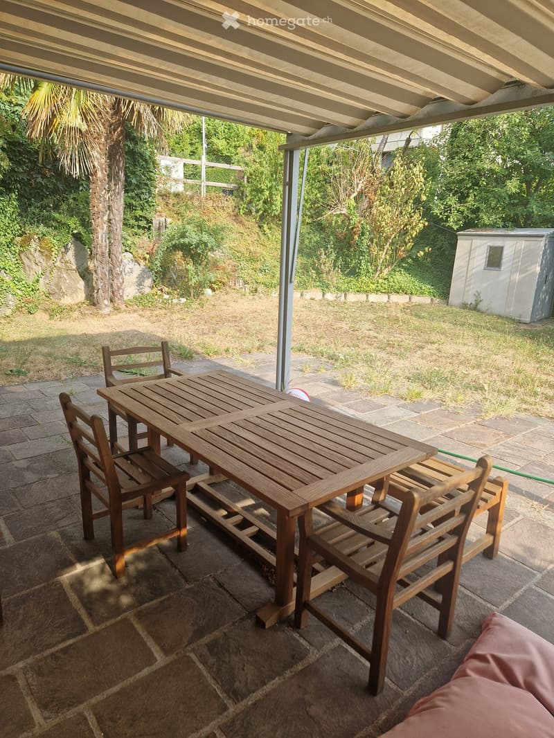 Wooden table and chairs on a stone patio under a covered area, surrounded by greenery and a small shed.