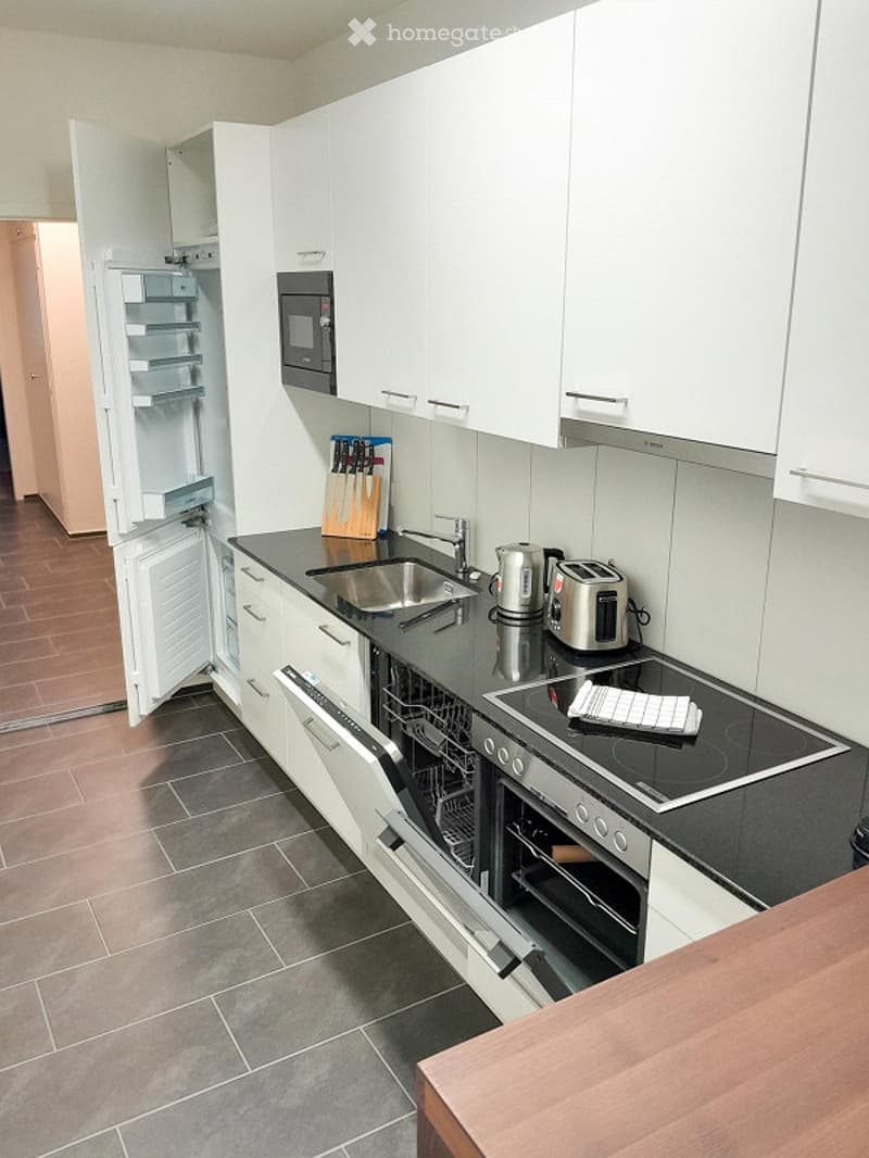 Modern kitchen with white cabinets, stainless steel appliances, and a black countertop. Visible items include a refrigerator, microwave, dishwasher, oven, sink, faucet, toaster, electric kettle, and a knife block.