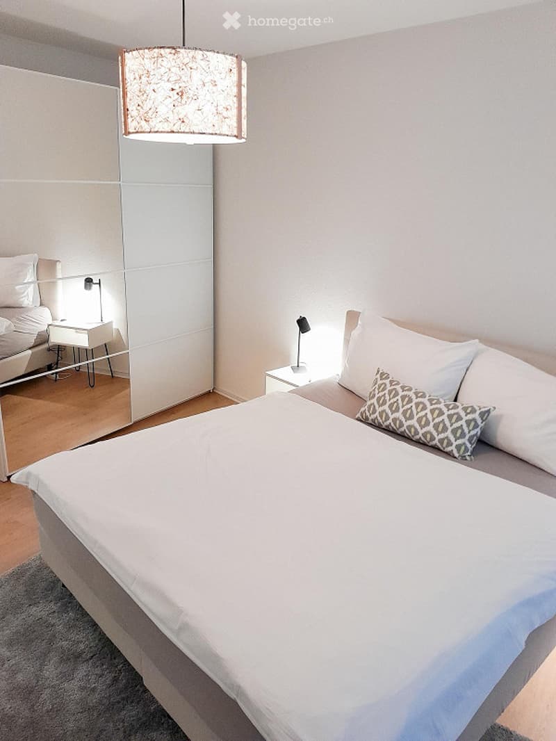 Modern bedroom with a large bed, white bedding, decorative pillows, nightstands with lamps, and a large mirrored wardrobe.