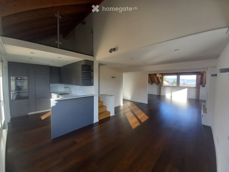 Open-plan space with a modern kitchen area featuring dark grey cabinets, a white countertop, and a sink. The main area has wooden flooring and large windows offering a scenic view. The ceiling is white with recessed lighting, and there is a small staircas