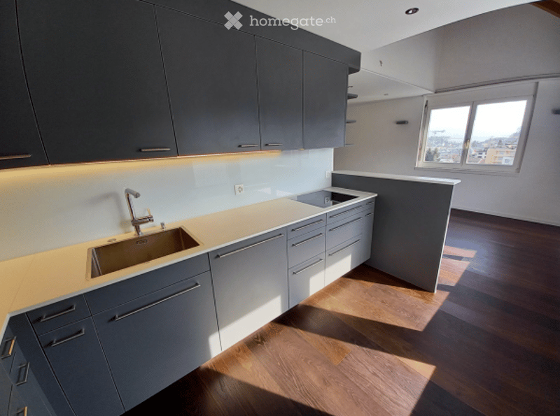 Modern kitchen with grey cabinets, wooden flooring, and a sink with a faucet. Natural light streams in from a window, casting shadows on the floor.