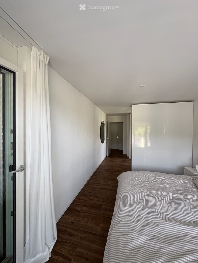 Modern bedroom with wooden flooring, white walls, a large bed with striped bedding, a white wardrobe, and a round mirror on the wall.