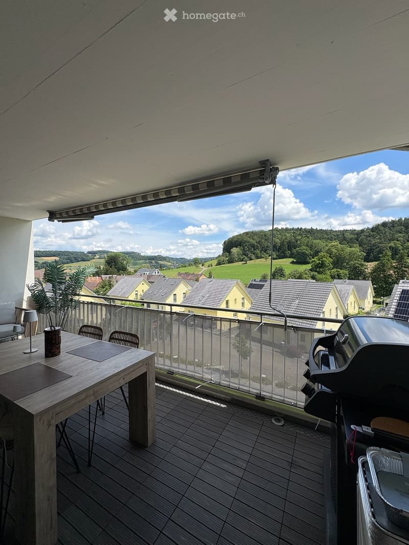 Balcony with wooden table, chairs, grill, and plant, overlooking a scenic countryside view.