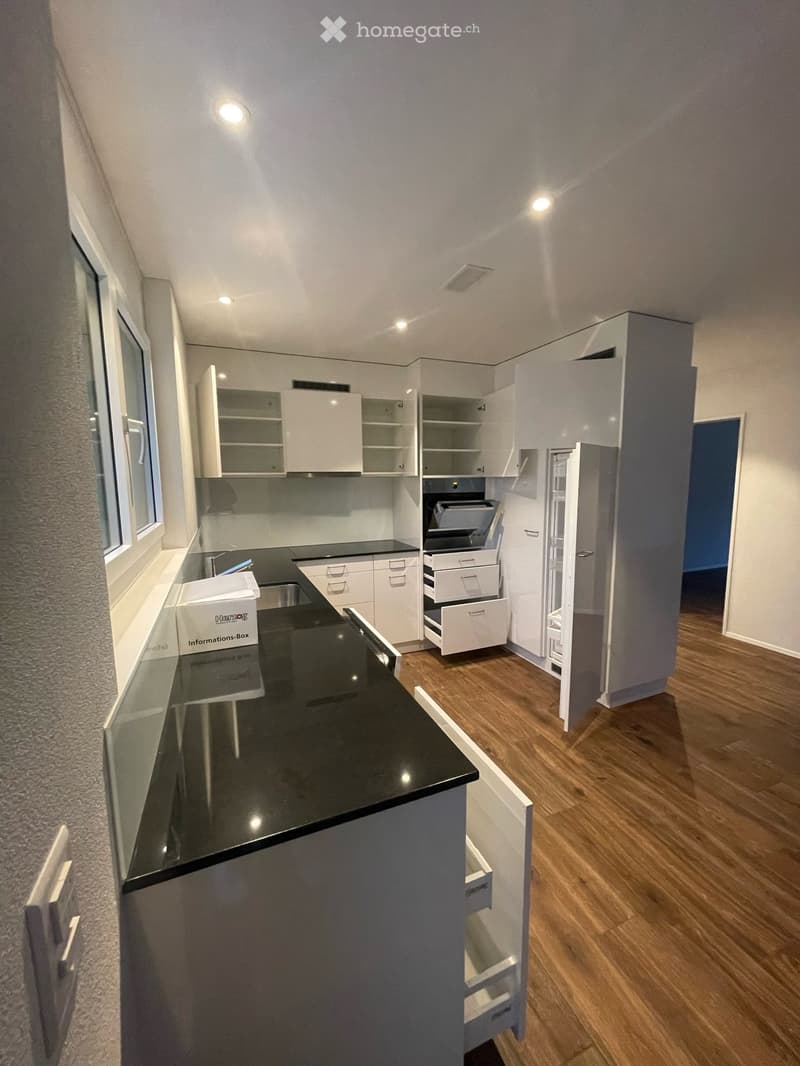 Modern kitchen with white cabinets, black countertop, stainless steel sink, and open shelving. Features include a refrigerator, oven, and wooden flooring.