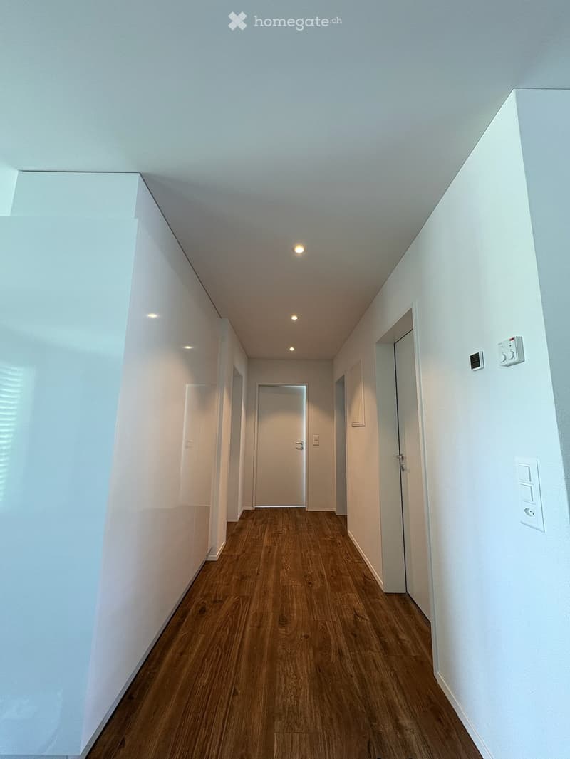 Clean, modern hallway with wooden flooring, white walls, and recessed lighting. Features several doors and electrical outlets.