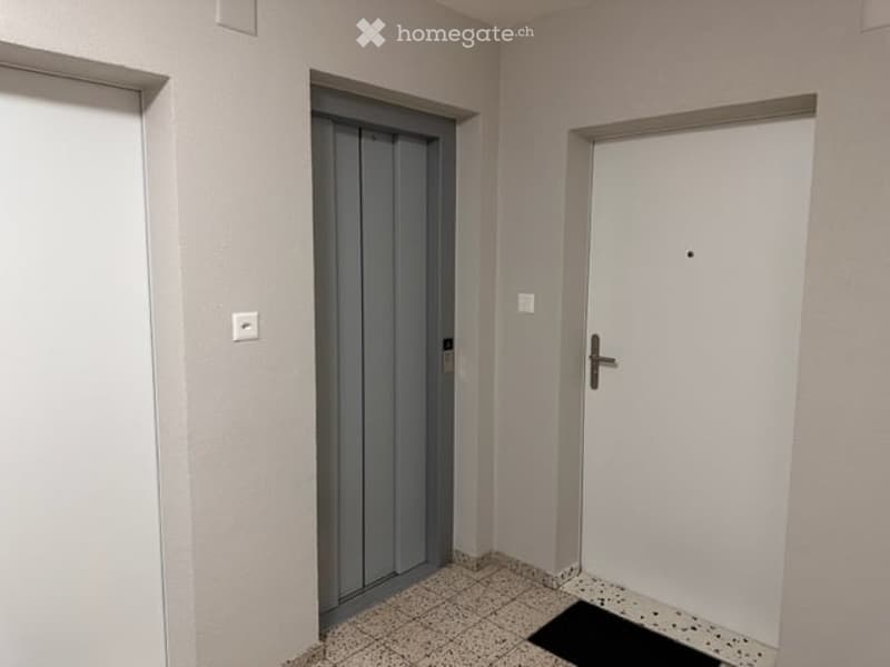 Empty hallway with tiled floor, white walls, and two doors: one grey elevator door and one white room door.