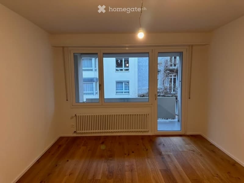 Empty room with wooden flooring, large windows, radiator, and sliding glass door leading to a small outdoor area.