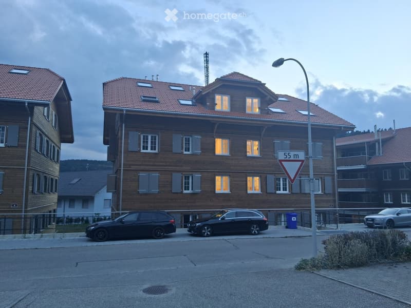 Multi-story wooden building with red-tiled roof, illuminated windows, and parked cars in front.