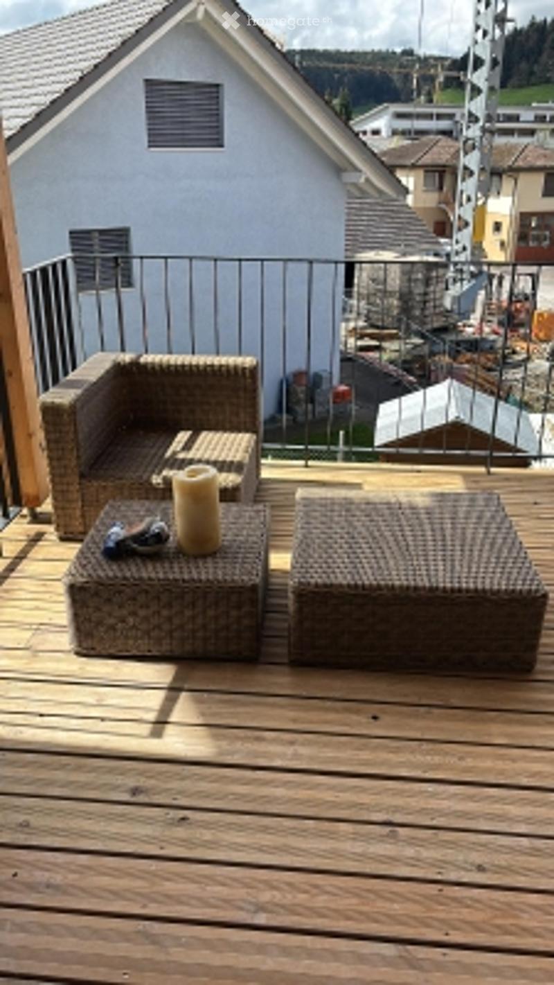 Wooden balcony with two wicker chairs and a small table, overlooking a residential area with houses and construction.