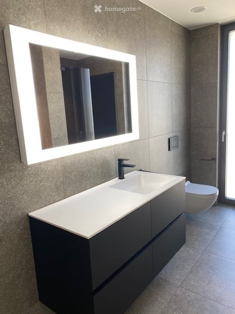 Modern bathroom with a sleek black vanity, white countertop, illuminated mirror, and a toilet visible in the background.
