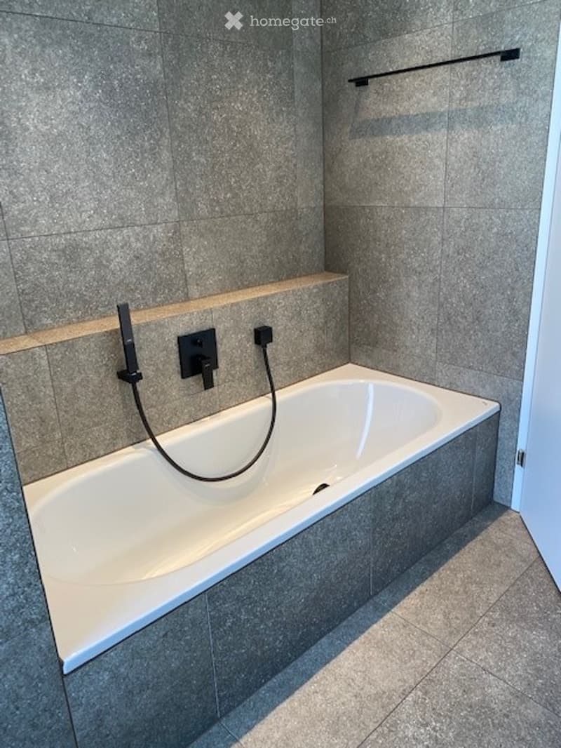 Modern bathroom with a white bathtub, grey tiled walls and floor, and a handheld showerhead attached to the wall.