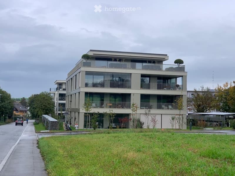 Modern multi-story residential building with large glass windows and balconies, surrounded by greenery and a paved walkway.