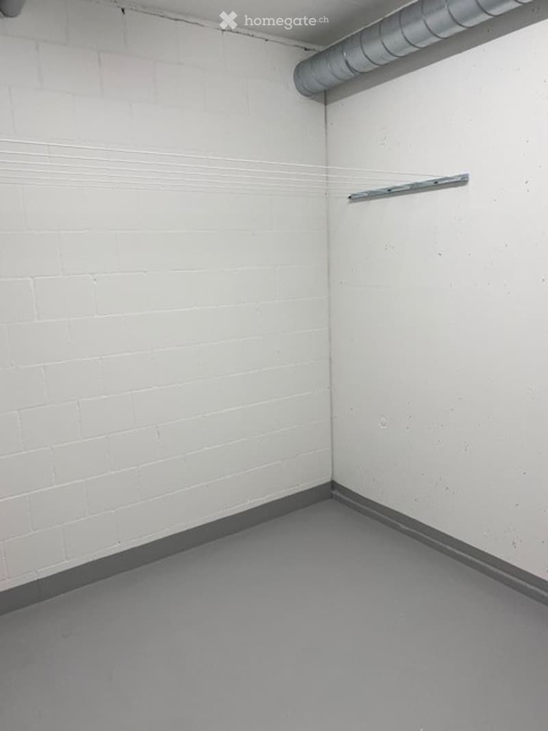 Empty room with white brick walls and a grey concrete floor, featuring a metal shelf on one wall and a visible pipe along the ceiling.