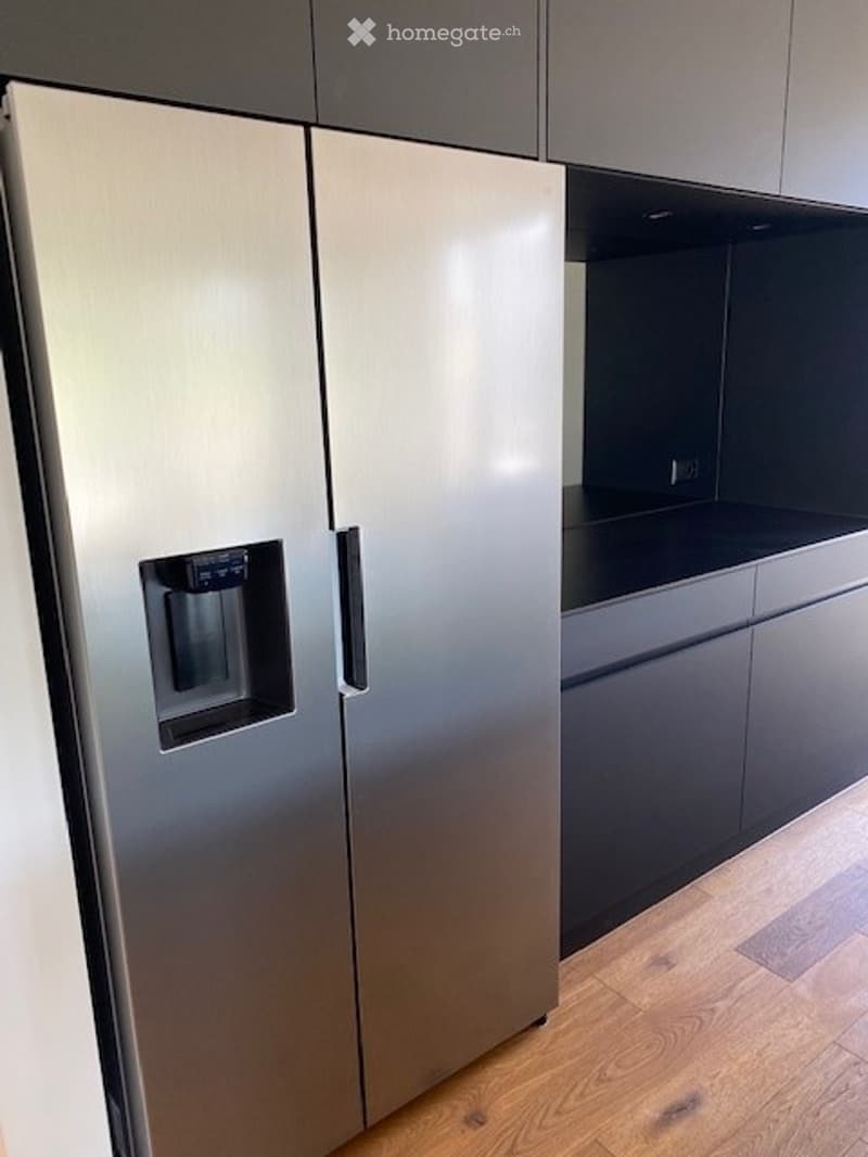 Modern kitchen with sleek, stainless steel refrigerator and dark grey cabinets, light wooden flooring.
