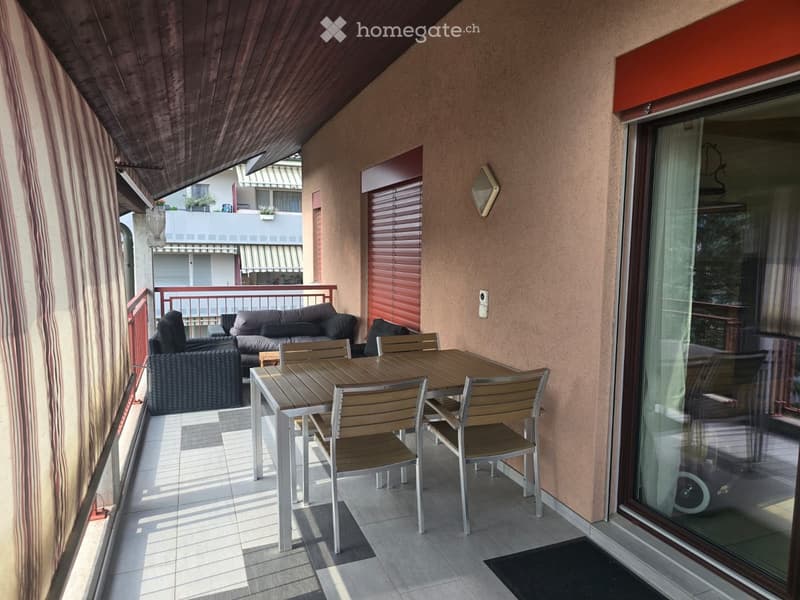 Balcony with a sofa, a wooden table, and chairs, featuring a wooden ceiling and a view of neighboring buildings.