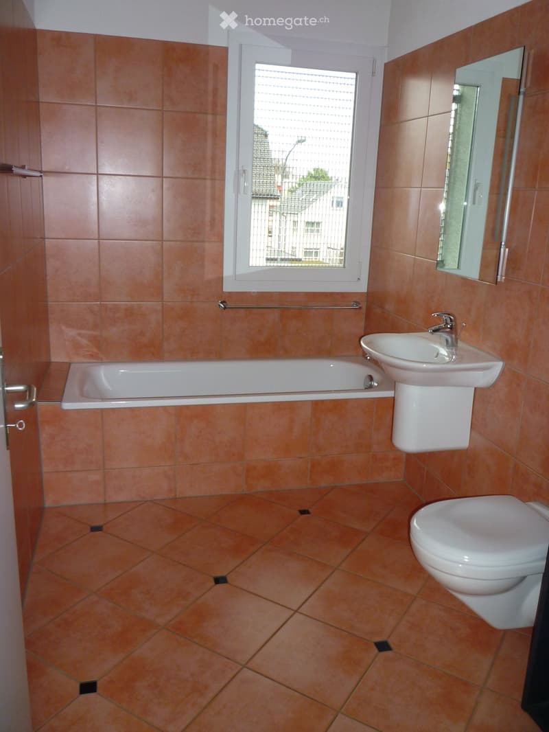 Bathroom with orange tiled walls and floor, featuring a bathtub, sink, toilet, and mirror. A window with blinds offers natural light.