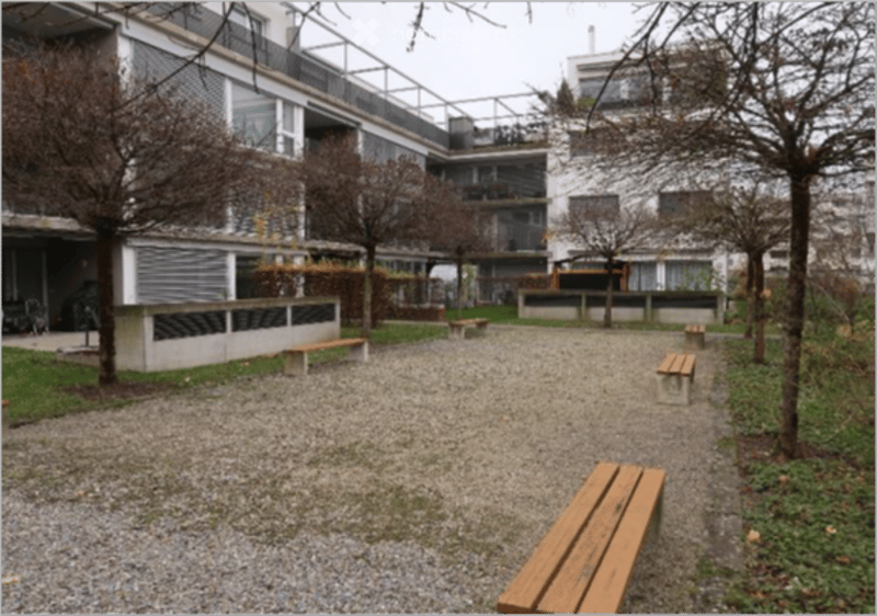 Courtyard area with gravel ground, several wooden benches, trees, and a modern building in the background.