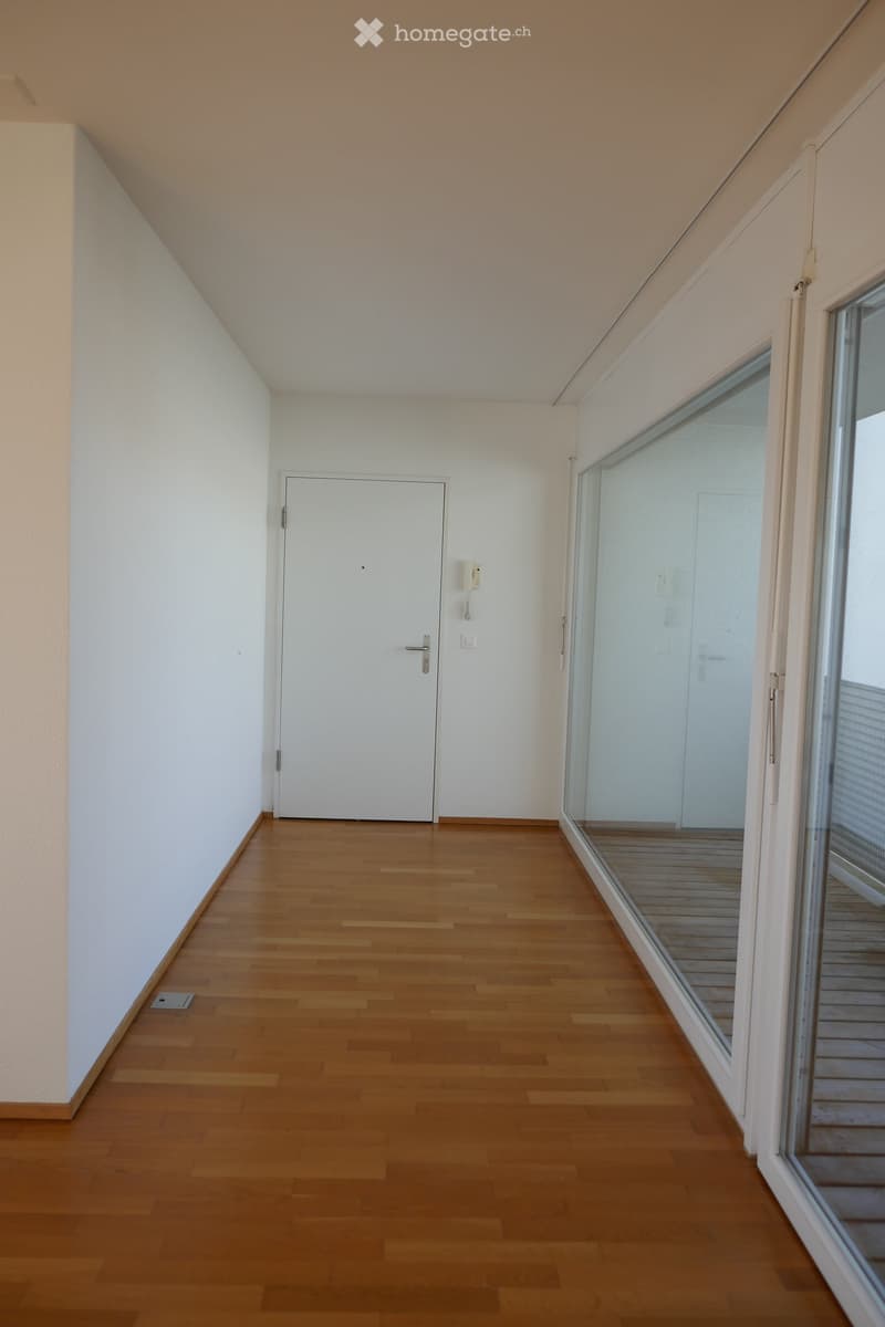 Empty hallway with wooden flooring, white walls, a closed white door, and a large glass sliding door leading to a balcony.