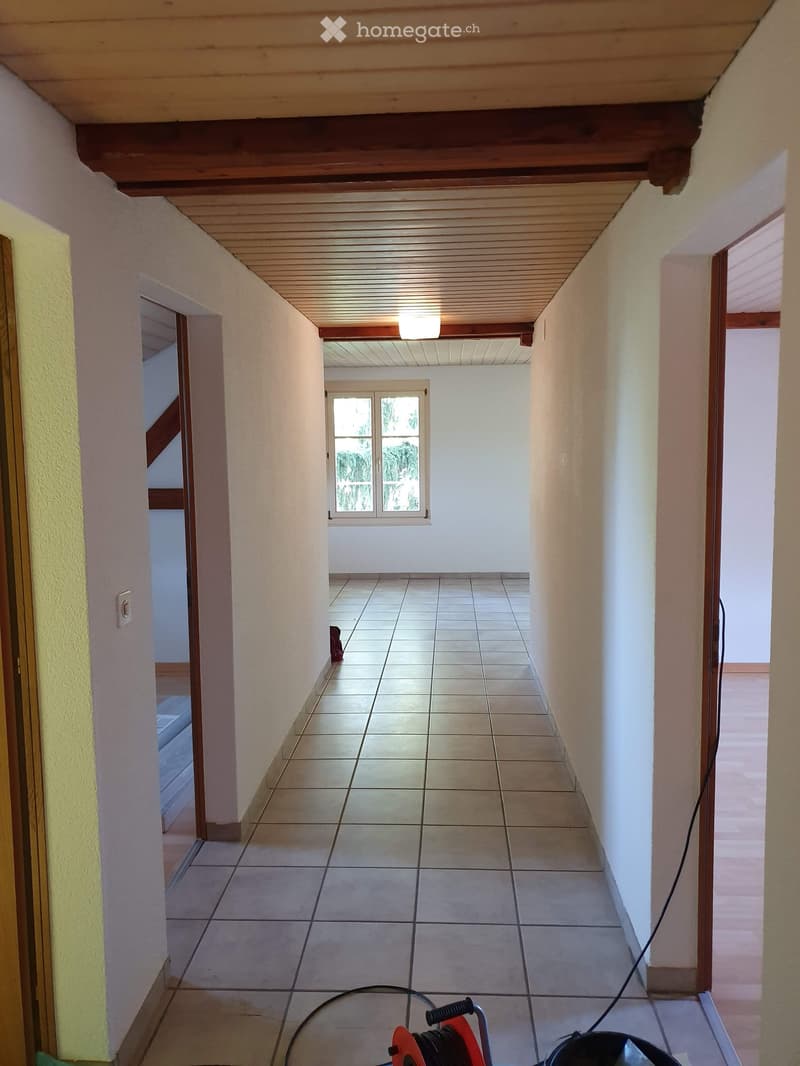 A long, narrow hallway with tiled flooring, white walls, and a wooden ceiling. There is a window at the far end, and the hallway is well-lit with a ceiling light fixture.