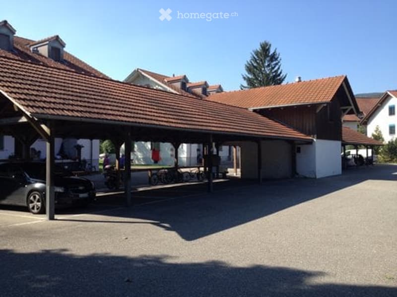 Car parked under a covered parking area, bicycles parked, building with brown roof, white walls, and a tree in the background.