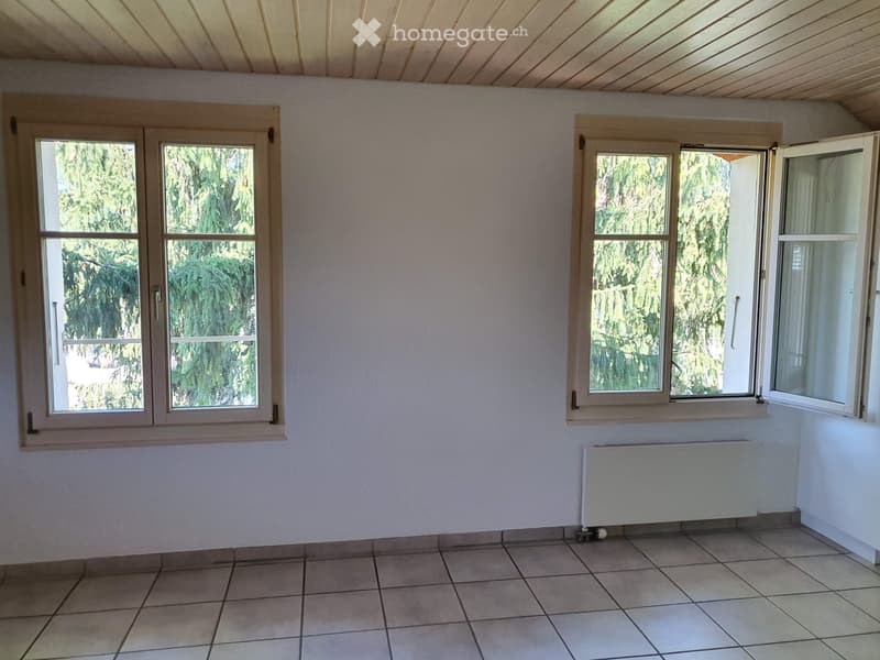 Empty room with tiled floor, white walls, and large windows with wooden frames. A radiator is visible on the right side.