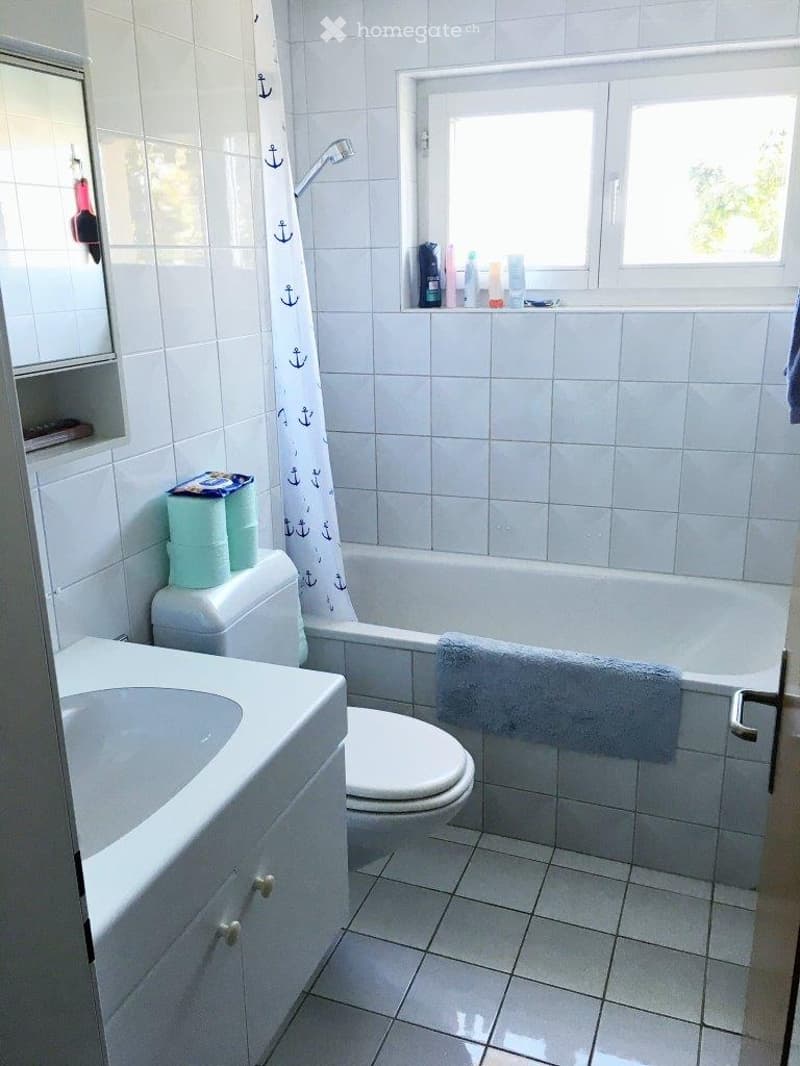 White bathroom with white tiles, bathtub, toilet, sink, shower curtain with anchor design