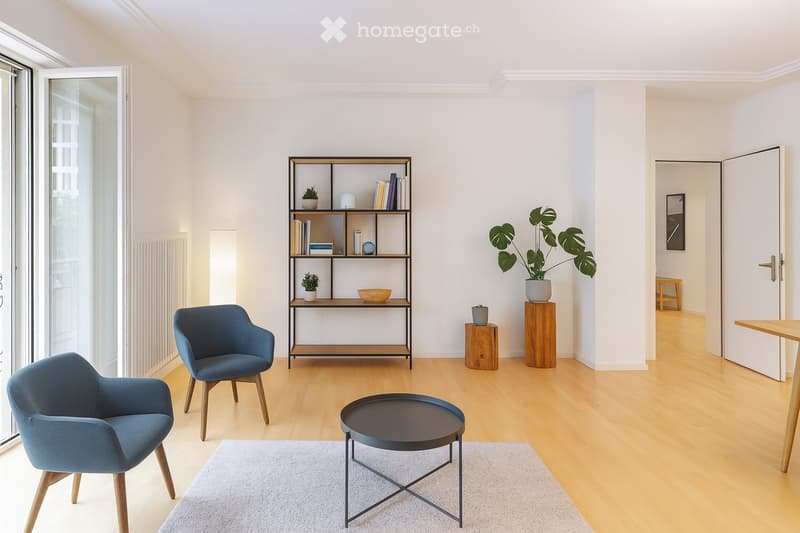 Modern living room with light wooden flooring, white walls, and large windows. Features include a grey sofa, two armchairs, a round coffee table, a bookshelf with books and decorative items, and a potted plant on a wooden stand.