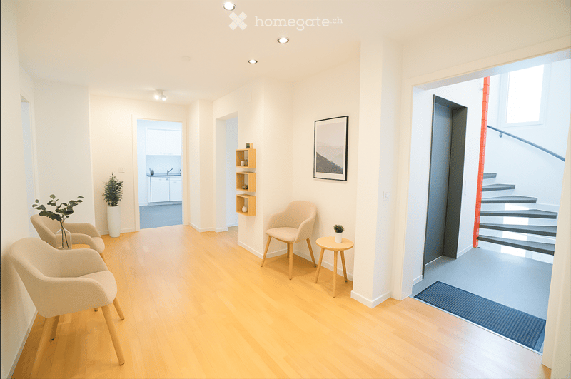 Bright, modern hallway with light wooden flooring, white walls, and recessed lighting. Features include a small seating area with chairs and a stool, a small table with a plant, a shelf with decorative items, and a framed picture on the wall. A staircase