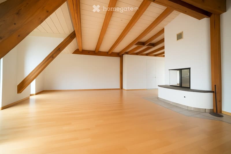 An empty room with wooden flooring, white walls, and exposed wooden beams on the ceiling. A small fireplace is visible on the right side.