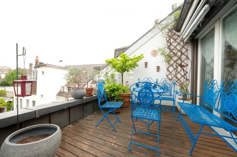 A wooden balcony with blue metal chairs and a table, surrounded by potted plants and a lantern.