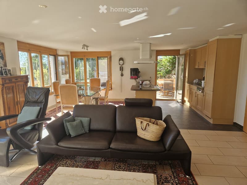 Bright living and dining area with large windows, leather sofa, coffee table, dining table with chairs, kitchen island, and cabinets.