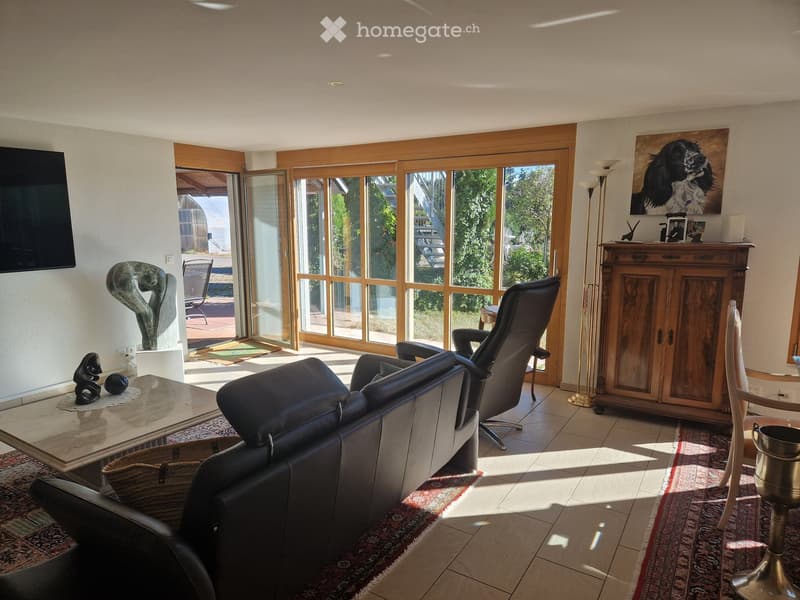 Bright living room with large windows and sliding glass doors leading to a patio. Features a leather sofa, armchair, coffee table, floor lamp, wooden cabinet, and artwork.