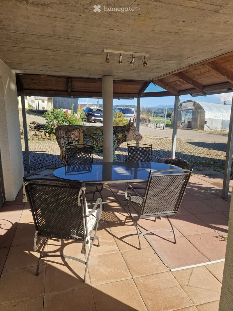Covered outdoor patio with a round table and four chairs, surrounded by a wooden ceiling and open sides, with a scenic view of the countryside.