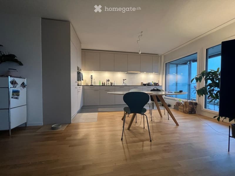 Modern kitchen and dining area with light wood flooring, white cabinets, and a central dining table with chairs. The kitchen features a refrigerator, sink, and various small appliances on the counter.
