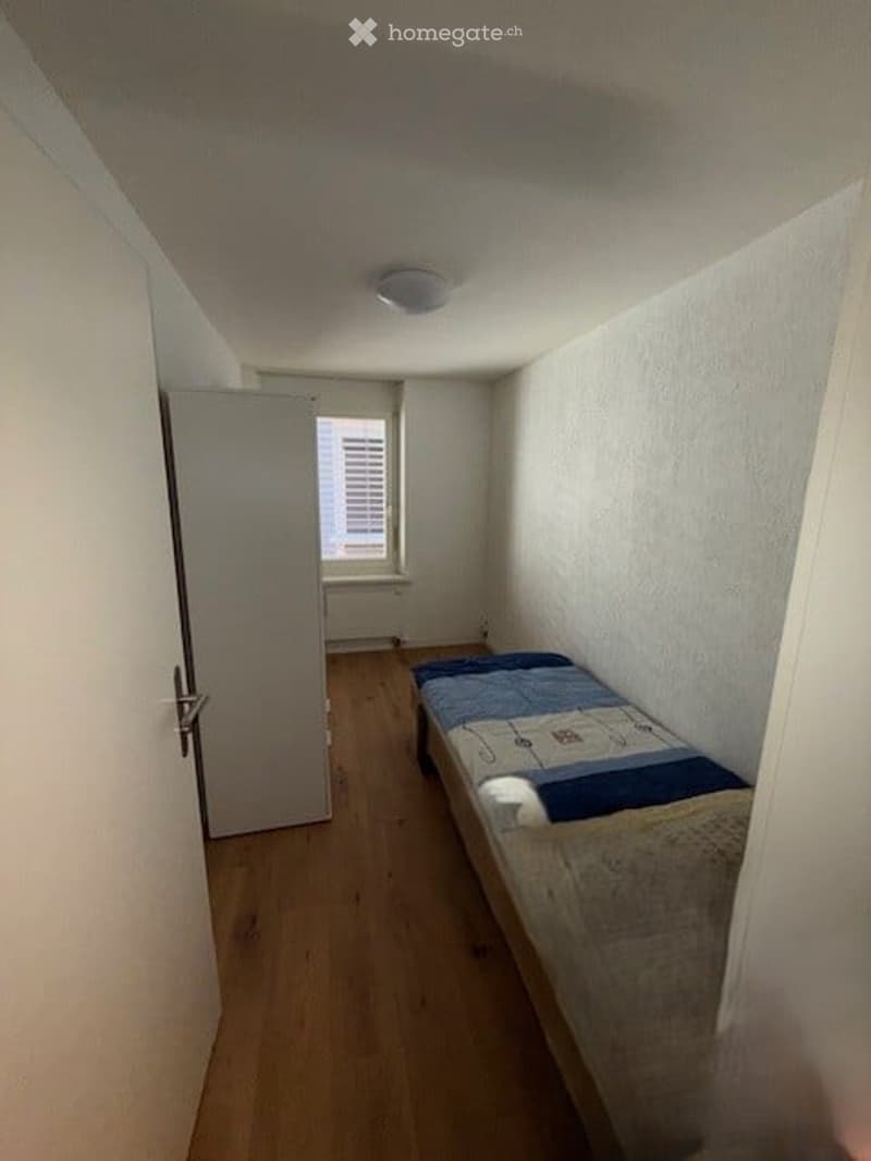 Small bedroom with wooden flooring, a single bed with blue and white bedding, and a wardrobe. The room has white walls and a window with blinds.