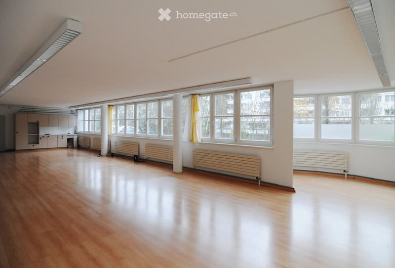 Empty room with wooden flooring, white walls, large windows with yellow curtains, radiators, and a small kitchen area with cabinets and a sink.