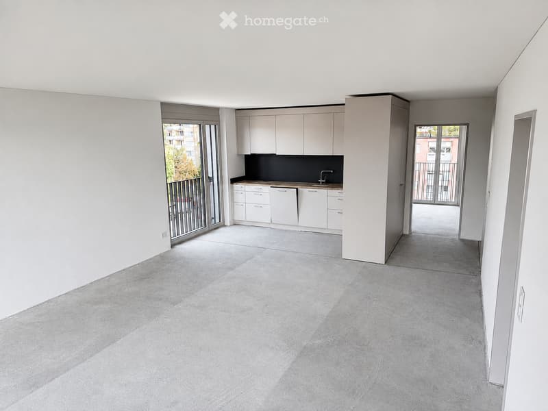 Empty room with white walls, grey concrete floor, and a small kitchen area with white cabinets and a sink.