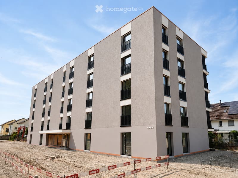 Modern multi-story residential building with light-colored facade, large windows, and balconies. The ground floor has an entrance area, and the building is surrounded by construction barriers and gravel.