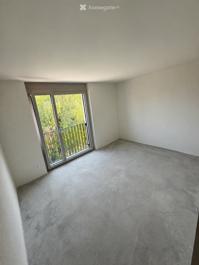 Empty room with smooth concrete floor and white walls, featuring a large sliding glass door leading to a balcony with metal railing and greenery visible outside.
