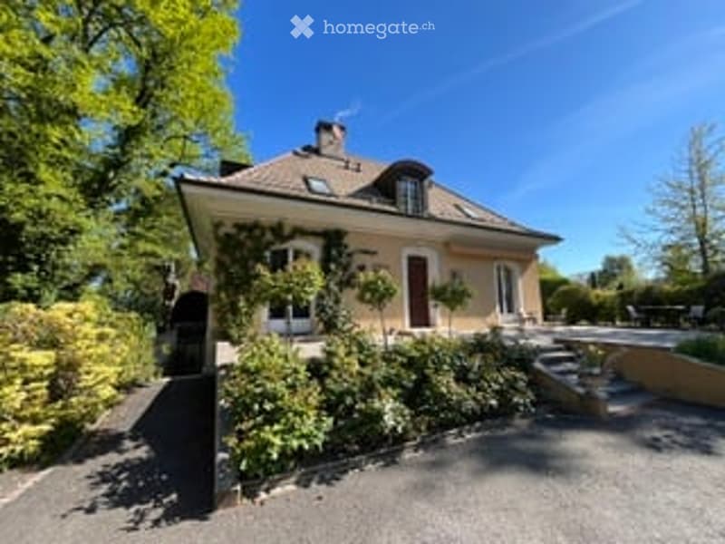 A two-story house with a beige exterior, multiple windows, and a well-maintained garden. The house features a sloped roof with dormer windows and a small staircase leading to the entrance.