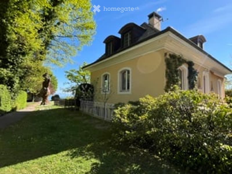 A charming yellow house with white-framed windows and a dark roof, surrounded by a well-maintained garden with lush greenery and neatly trimmed hedges.